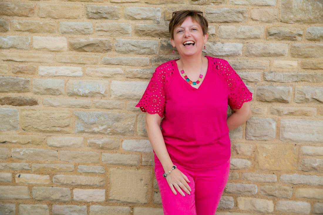 Kathryn (a white middle aged woman with brown hair and brown eyes) wearing a pink top and trousers leaning against a Cotswold stone wall smiling