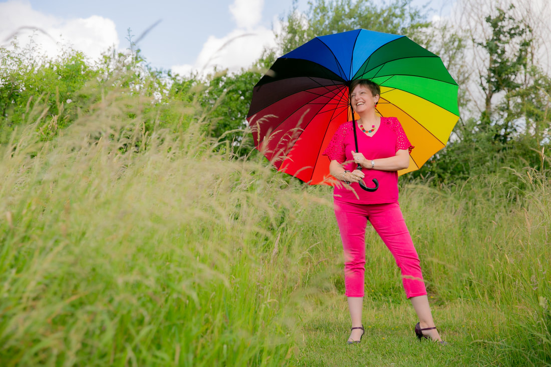 Kathryn (a white middle aged woman with brown hair and brown eyes) stood in a grassy field wearing a pink top and pink trousers holding a multi coloured umbrella